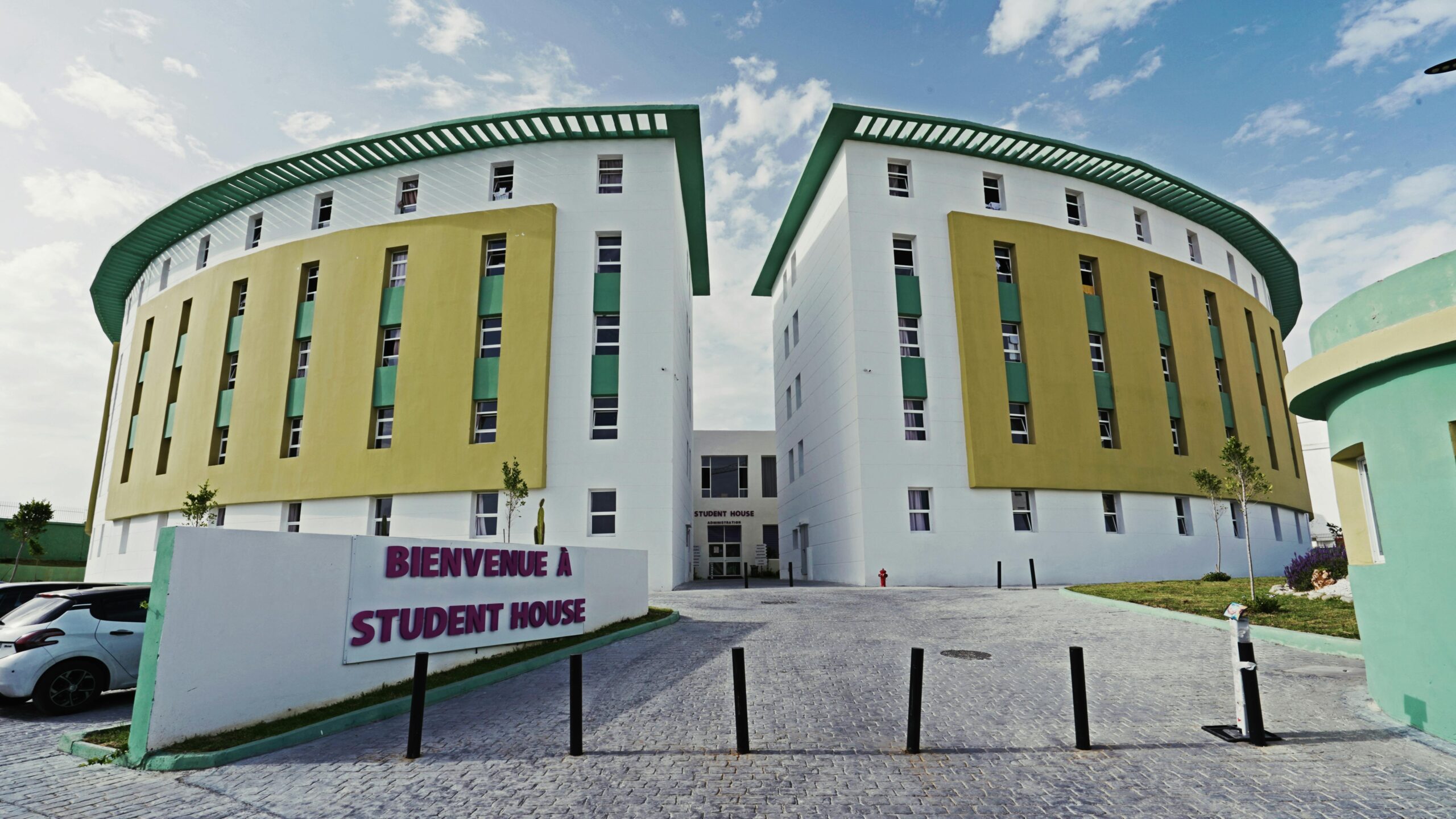 A modern student housing building with a welcoming sign that reads "Bienvenue à Student House," featuring contemporary architecture with white, yellow, and green accents.