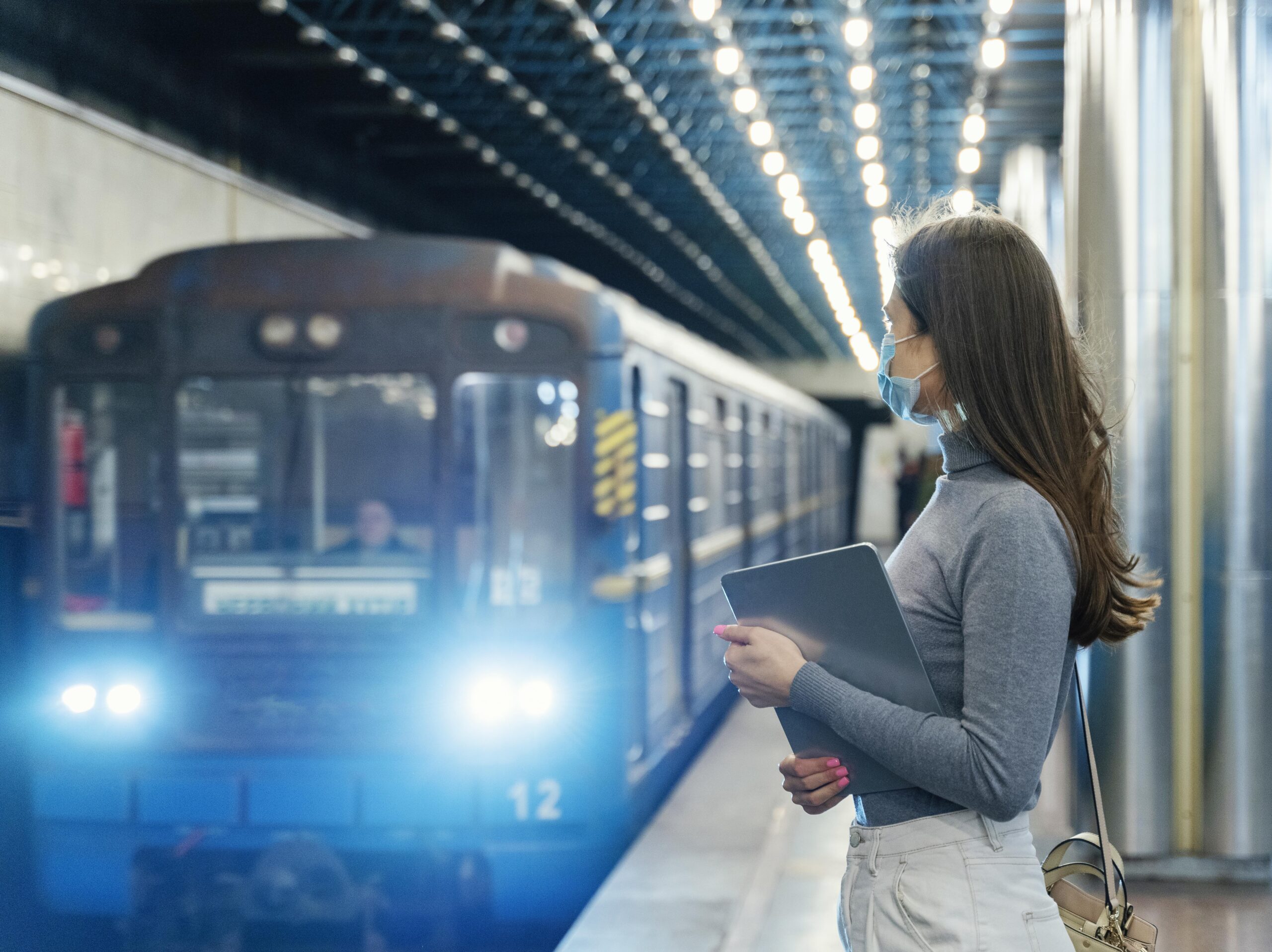 Young woman wearing a face mask waiting at a subway station with a tablet in hand, as a blue train arrives, representing public transport in New Zealand.