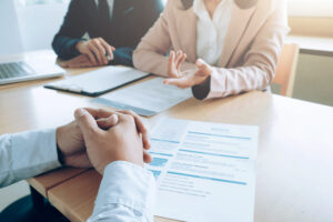 A professional setting featuring a UK job interview, where a candidate sits across from two interviewers discussing a resume. The scene includes documents, a laptop, and business attire, emphasizing a formal recruitment process.