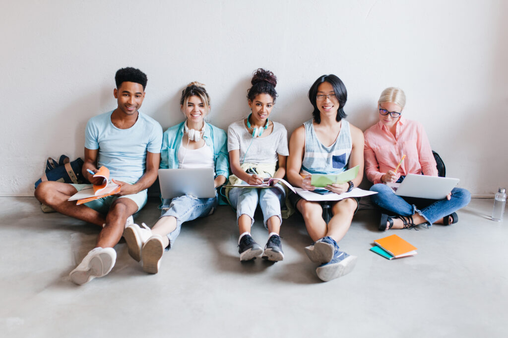 A group of diverse students sitting on the floor, using laptops and notebooks, symbolizing collaboration and the importance of staying connected in education and social life.