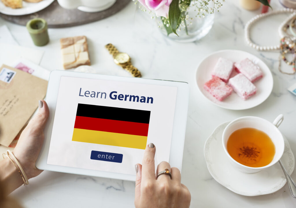 A person holding a tablet displaying "Learn German" with a German flag and an enter button on the screen. The tablet is placed on a white marble table with a cup of tea, pink pastries, a gold watch, letters, and pearl jewelry nearby.