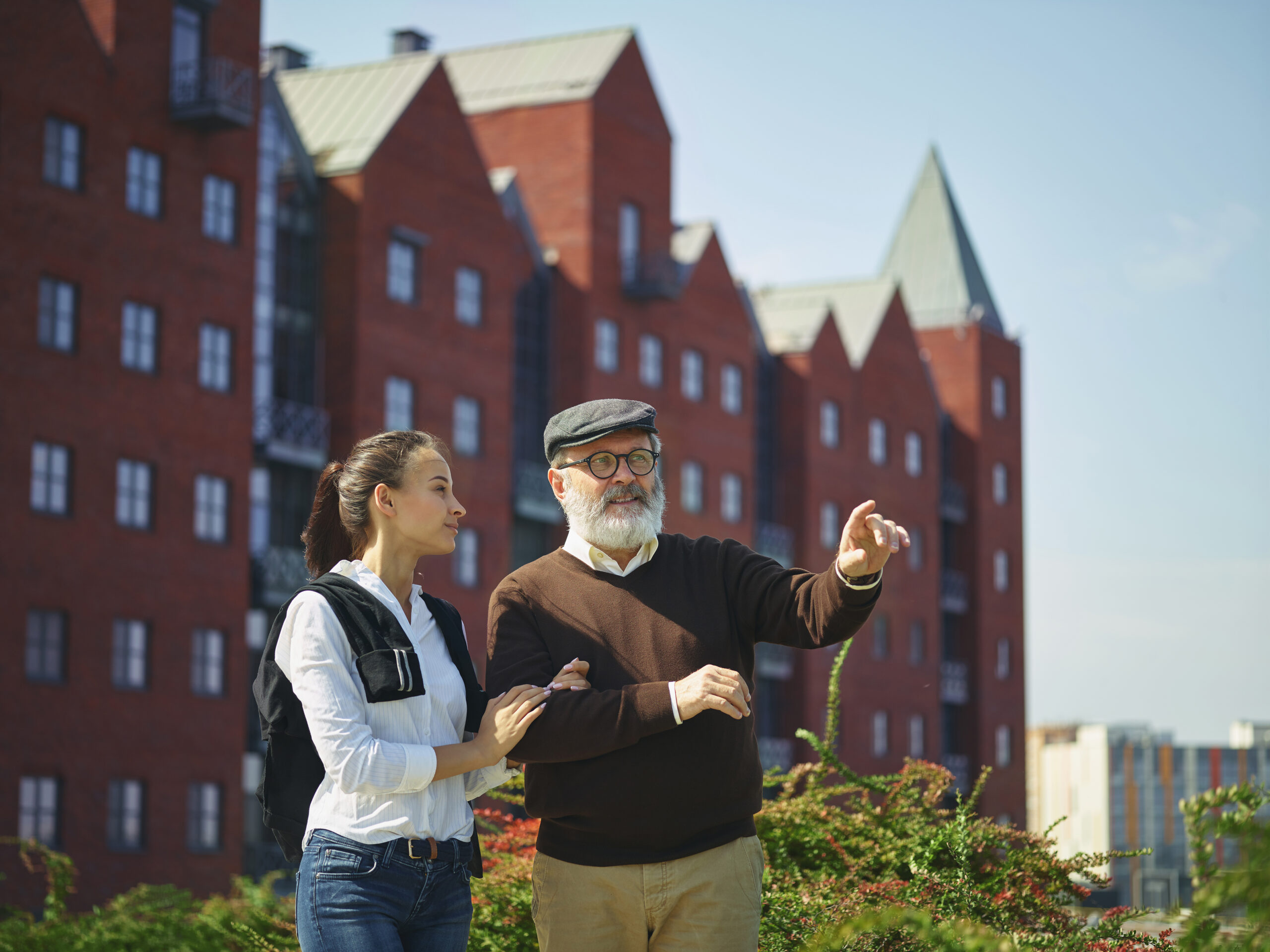 young woman and an elderly man stand together outdoors in Denmark, with the man pointing towards something in the distance. The young woman, dressed in casual attire with a backpack, listens attentively, symbolizing guidance and learning. The background features modern red-brick buildings with steep roofs, reminiscent of Danish architecture. This image represents the concept of Child's Education in Denmark, emphasizing mentorship, intergenerational learning, and knowledge-sharing.