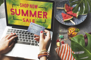 A person shopping online using a laptop displaying a "Summer Sale" promotion, holding a credit card for payment. The scene includes tropical elements, snacks, and a refreshing drink, representing student discounts & deals on seasonal purchases.