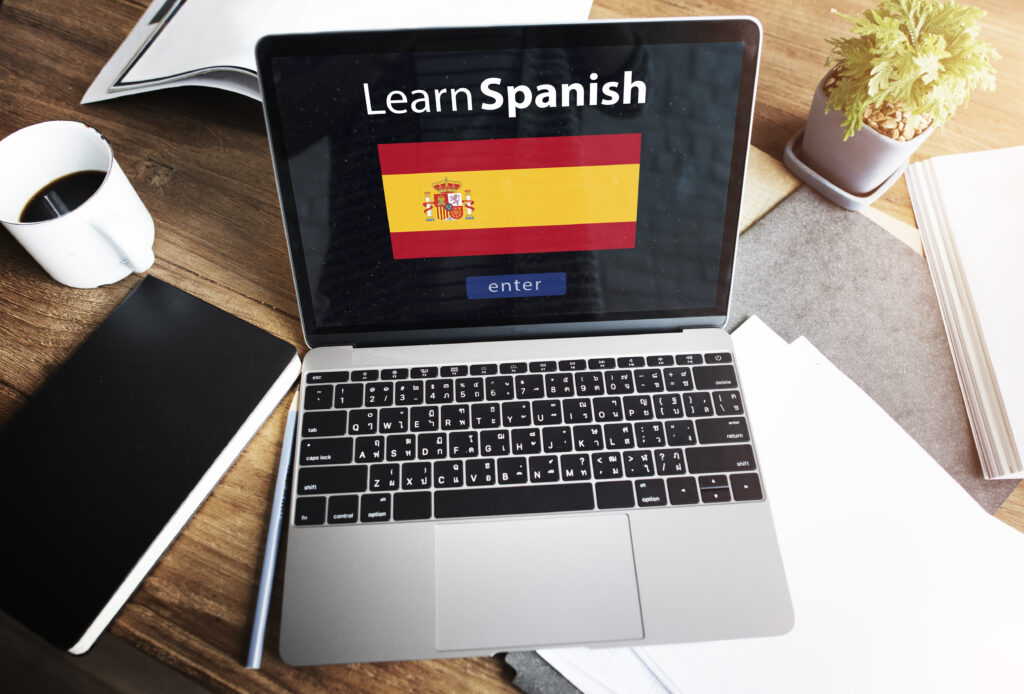 Open laptop displaying 'Learn Spanish' with the Spanish flag on the screen, symbolizing Spanish education through online learning. The workspace includes a coffee cup, notebook, and plant, creating a productive study environment.