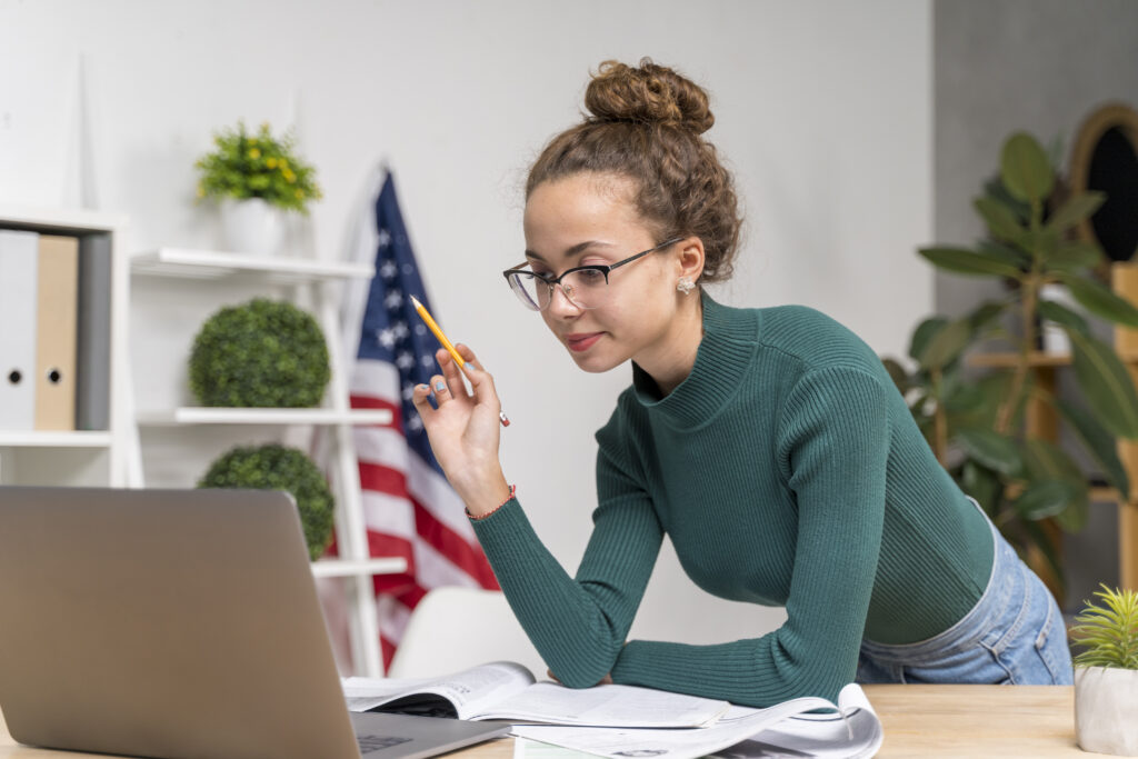 oung woman researching jobs and work placement in the UK on a laptop, surrounded by study materials, with an American flag in the background.