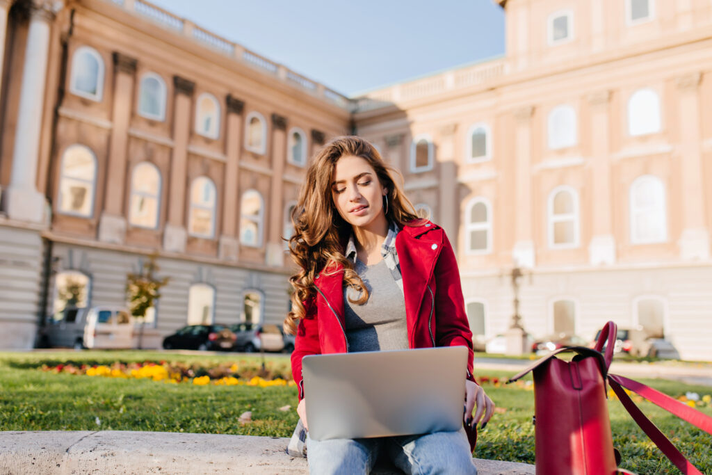 Young woman sitting outdoors with a laptop in front of a historic university building, representing security in Australia. She appears focused, symbolizing a safe and comfortable study environment. A red handbag rests beside her on the grass, adding to the relaxed yet secure atmosphere.