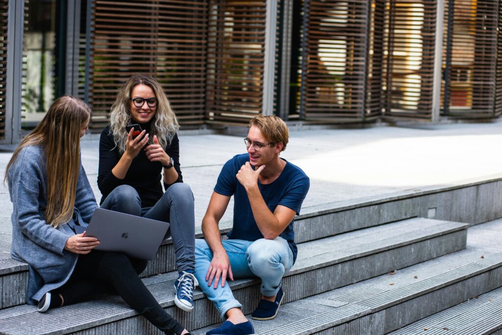 Three university students in Spain sitting on outdoor steps, engaged in a friendly conversation. One student is holding a smartphone while another has a laptop, suggesting a collaborative or study-related discussion. The background features modern architecture with wooden blinds, creating a vibrant campus atmosphere.
