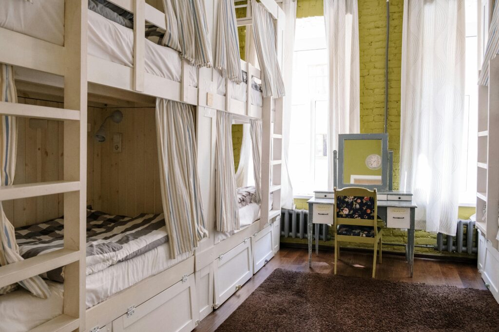 Cozy dormitory-style accommodation for international students featuring wooden bunk beds with privacy curtains, a vintage dressing table, and natural light streaming through large windows. The room offers a comfortable and affordable living space for students or travelers.