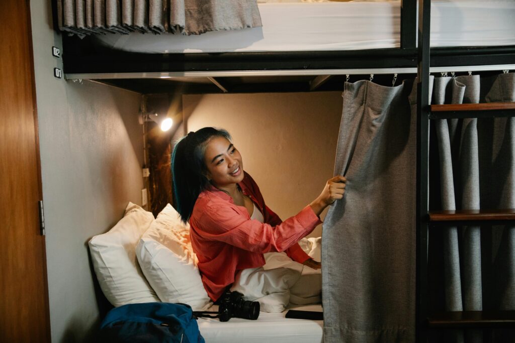 Young woman in a cozy hostel bunk bed, smiling while adjusting the privacy curtain, representing affordable student accommodation in Spain on a student budget