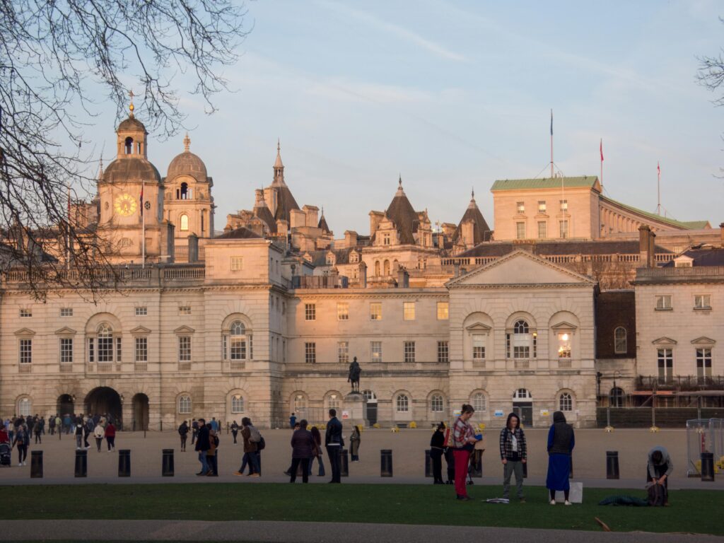 Social Life in the UK: A lively scene outside a historic building in London, with people strolling, chatting, and engaging in activities on a public square. The warm glow of the evening sun highlights the architectural beauty, capturing the essence of daily life and community interactions in the UK.