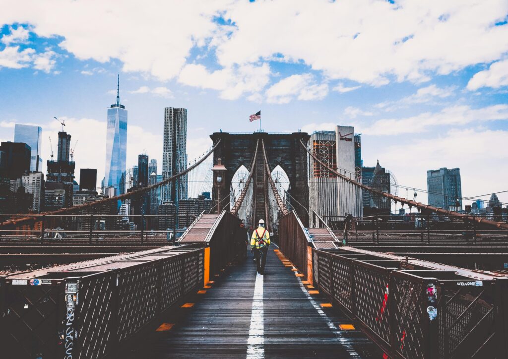 Staying Safe in the USA: A worker in a high-visibility vest walks along the Brooklyn Bridge with the New York City skyline in the background, symbolizing urban security and awareness in a bustling metropolis