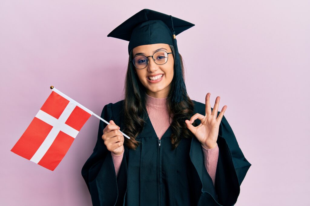 Smiling International Graduate wearing a black cap and gown, holding a Denmark flag and making an 'OK' hand gesture against a pink background.
