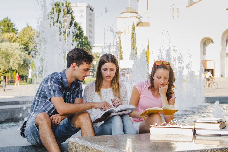 Three people sitting on the edge of a fountain outdoors, surrounded by books. They appear to be studying or discussing something together, with one person pointing at a book. The setting is sunny, with trees and a building visible in the background.