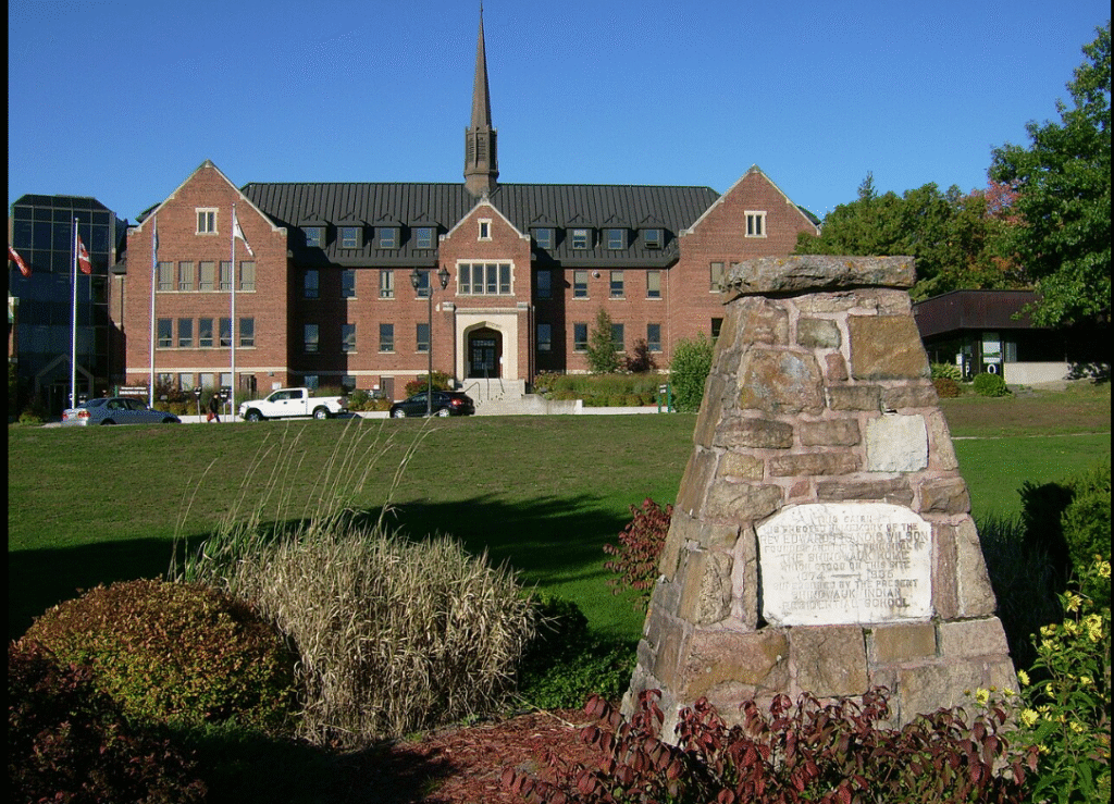 A large brick building at Algoma University with a central steeple and symmetrical architecture, set against a clear blue sky. In the foreground, there is a stone monument with an engraved plaque, surrounded by landscaped greenery and a well-maintained lawn.