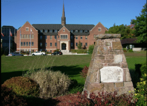 A large brick building at Algoma University with a central steeple and symmetrical architecture, set against a clear blue sky. In the foreground, there is a stone monument with an engraved plaque, surrounded by landscaped greenery and a well-maintained lawn.