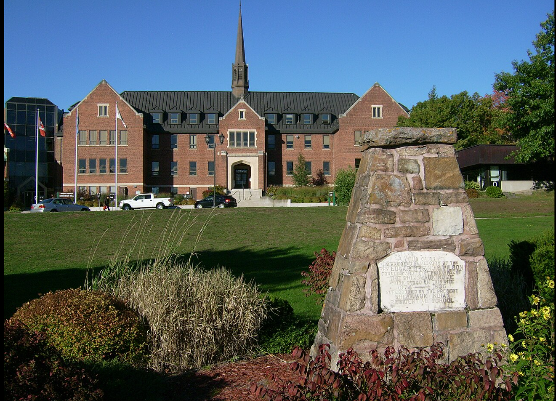 A large brick building at Algoma University with a central steeple and symmetrical architecture, set against a clear blue sky. In the foreground, there is a stone monument with an engraved plaque, surrounded by landscaped greenery and a well-maintained lawn.