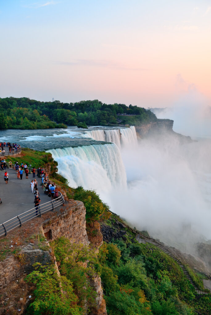 Tourists viewing the majestic Niagara Falls at dusk, with powerful waterfalls cascading over rocky cliffs surrounded by lush greenery and mist rising into the air.