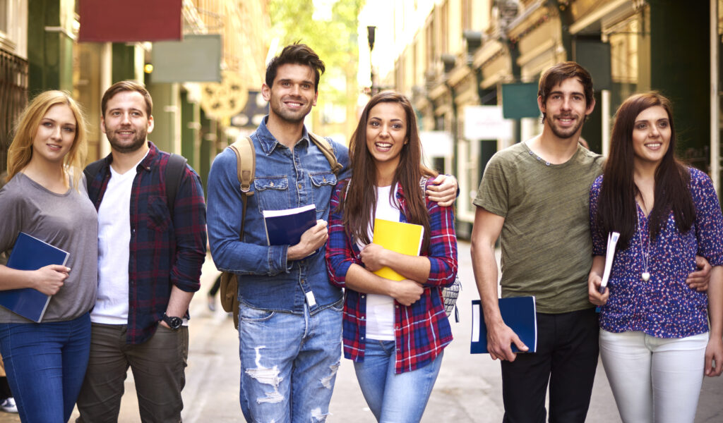 A group of six smiling college students, three men and three women, stand closely together on a city street holding notebooks. They appear cheerful and casually dressed, enjoying a sunny day, with shops and greenery visible in the background.