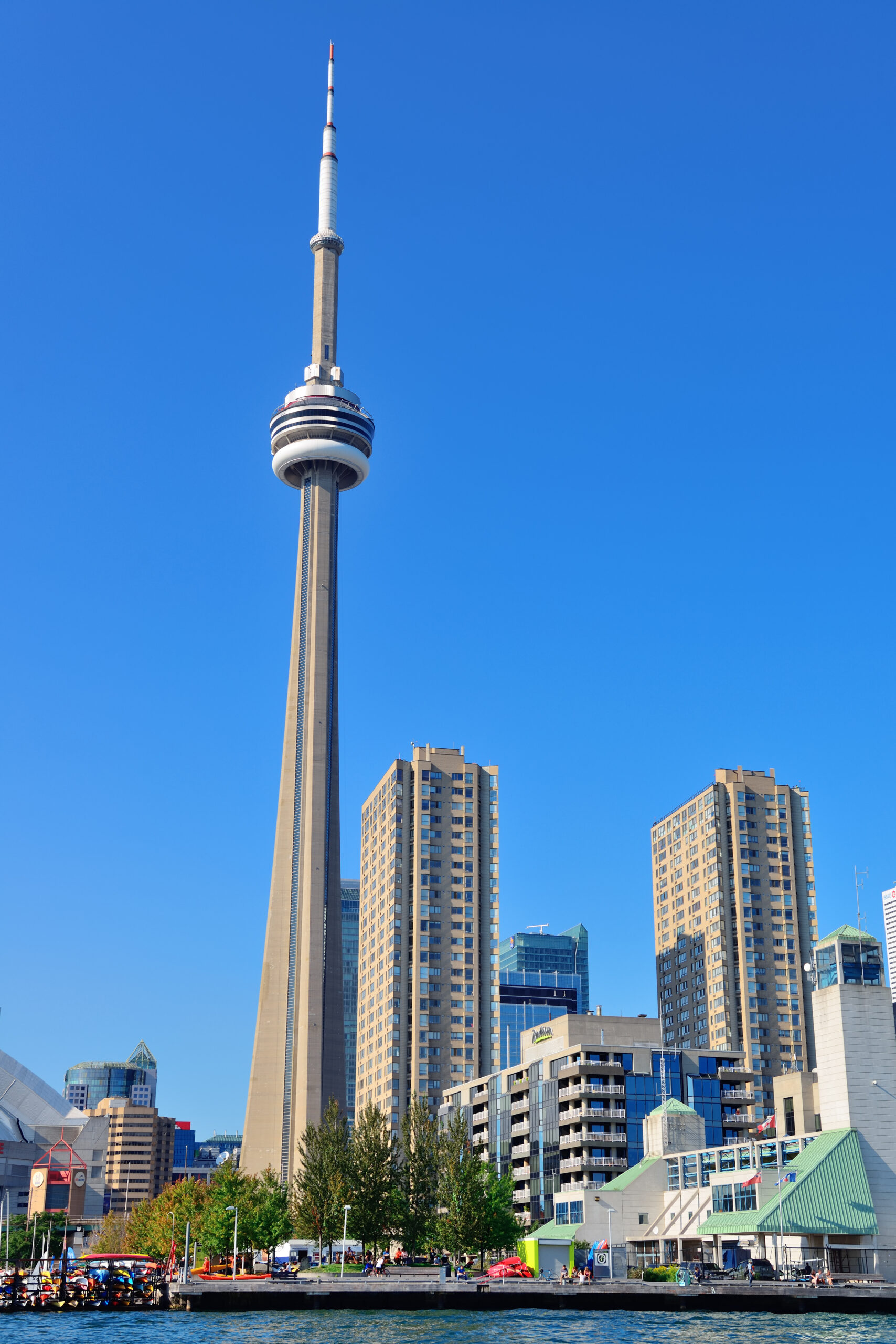 A clear daytime view of the CN Tower in Toronto, Canada, rising prominently above surrounding high-rise buildings. The image is taken from across the waterfront, with people and colorful kayaks visible along the harbor in the foreground under a bright blue sky.