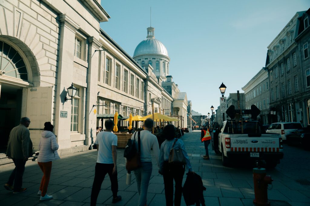 People stroll past a cafe on a cobblestone street in Old Montreal, with the iconic dome of a building in the background.