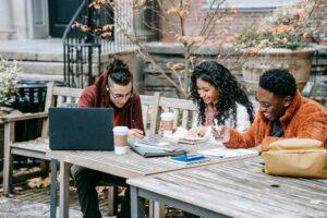 Three students collaborate on a study project outdoors at a wooden table.
