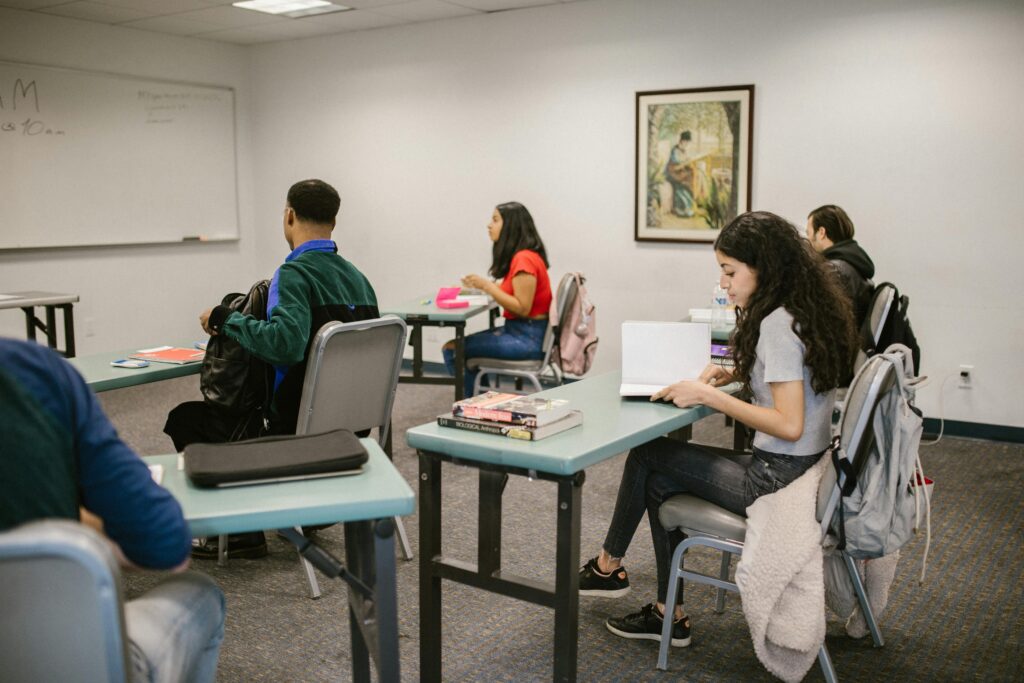 Students sit at desks in a classroom, some writing or reading, with notebooks and textbooks on their tables. A painting hangs on the wall and a whiteboard is visible.