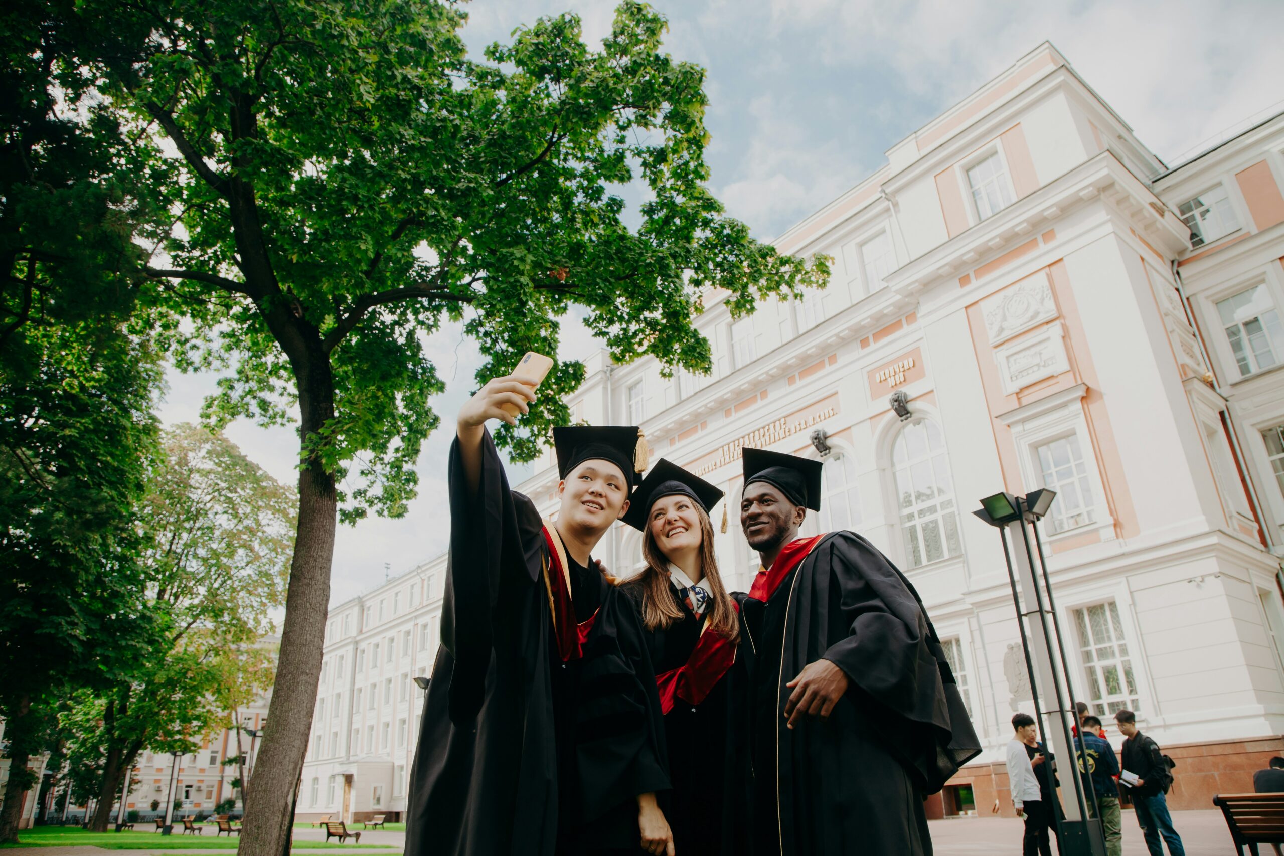 International computer science students in graduation gowns taking a selfie on a university campus, representing global co-op programs and international study opportunities for tech students.