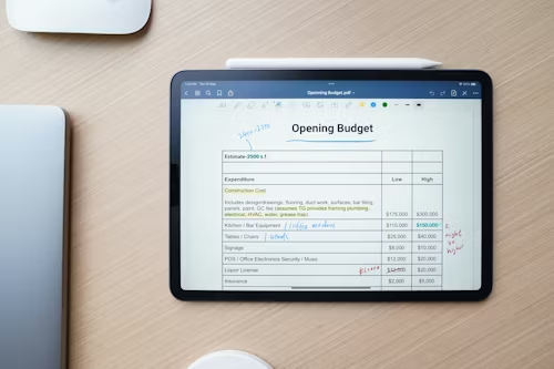 Tablet displaying an “Opening Budget” spreadsheet on a wooden desk, showing estimated expenses for construction, kitchen equipment, chairs, and licenses, with handwritten notes and a stylus nearby.