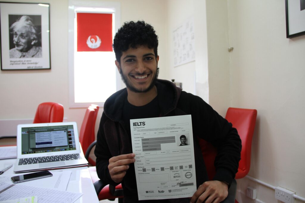 Smiling student sitting in an office and holding an IELTS test report form, with a laptop and red chairs in the background.
