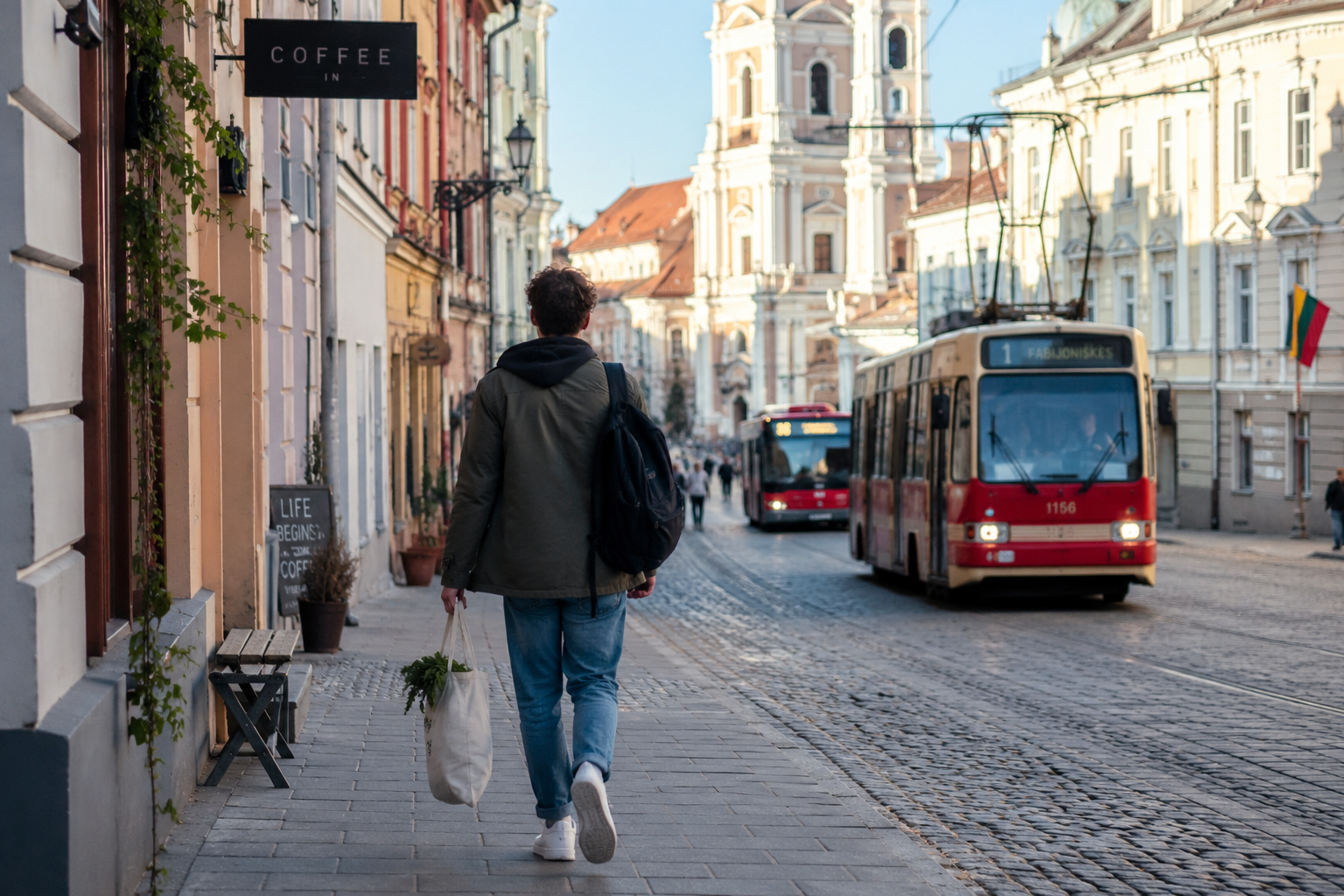 International student walking through Vilnius Old Town with a backpack and reusable grocery bag, surrounded by historic architecture, cobblestone streets, and public transportation, illustrating affordable student life in Lithuania.