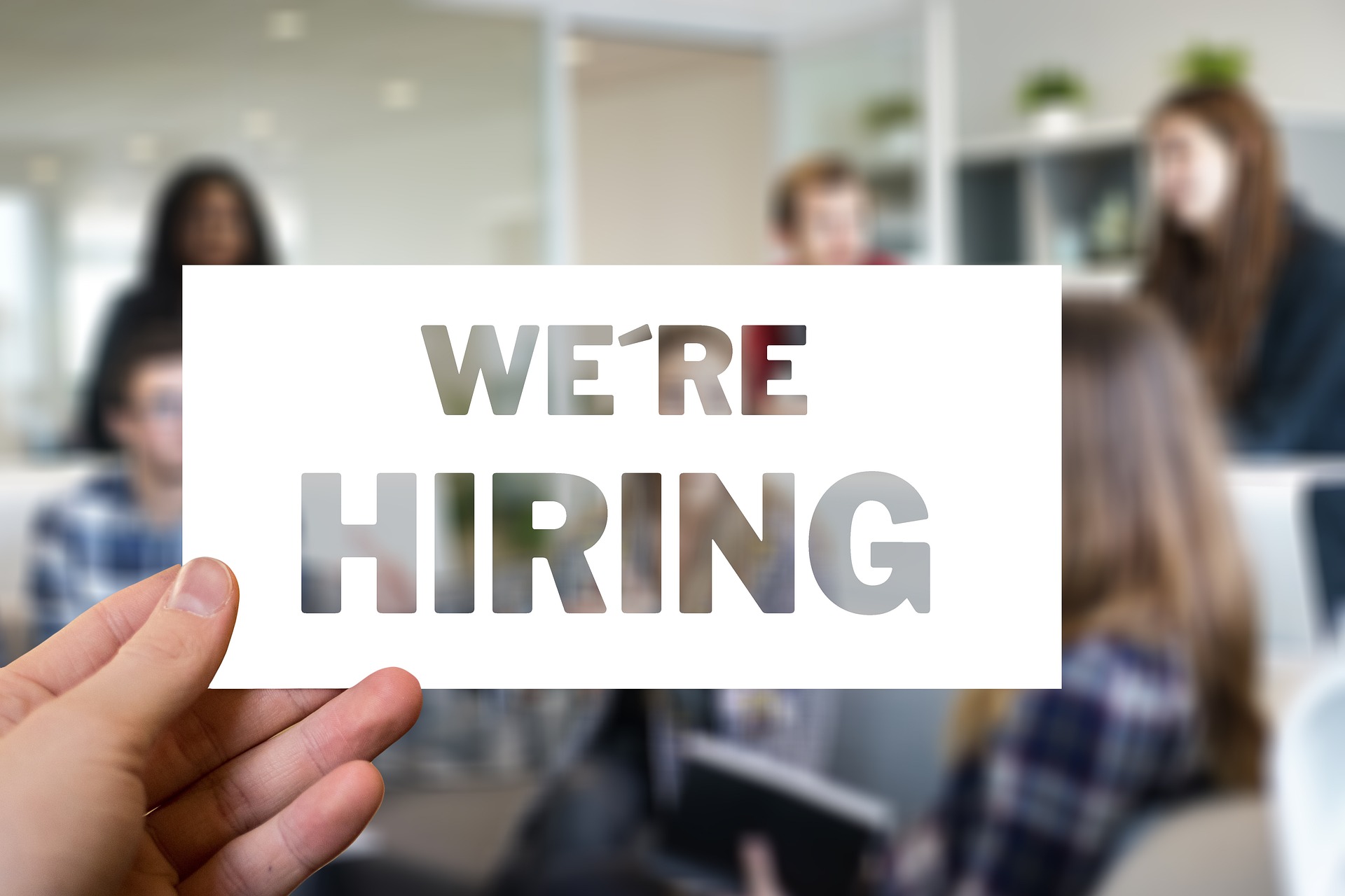 Person holding a card with the words “We’re Hiring” in front of a blurred office meeting, representing networking and access to unlisted international job opportunities in the hidden job market.