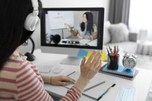 Student attending an online international degree class from home, participating in a live virtual lecture with headphones, computer, and study materials on the desk.