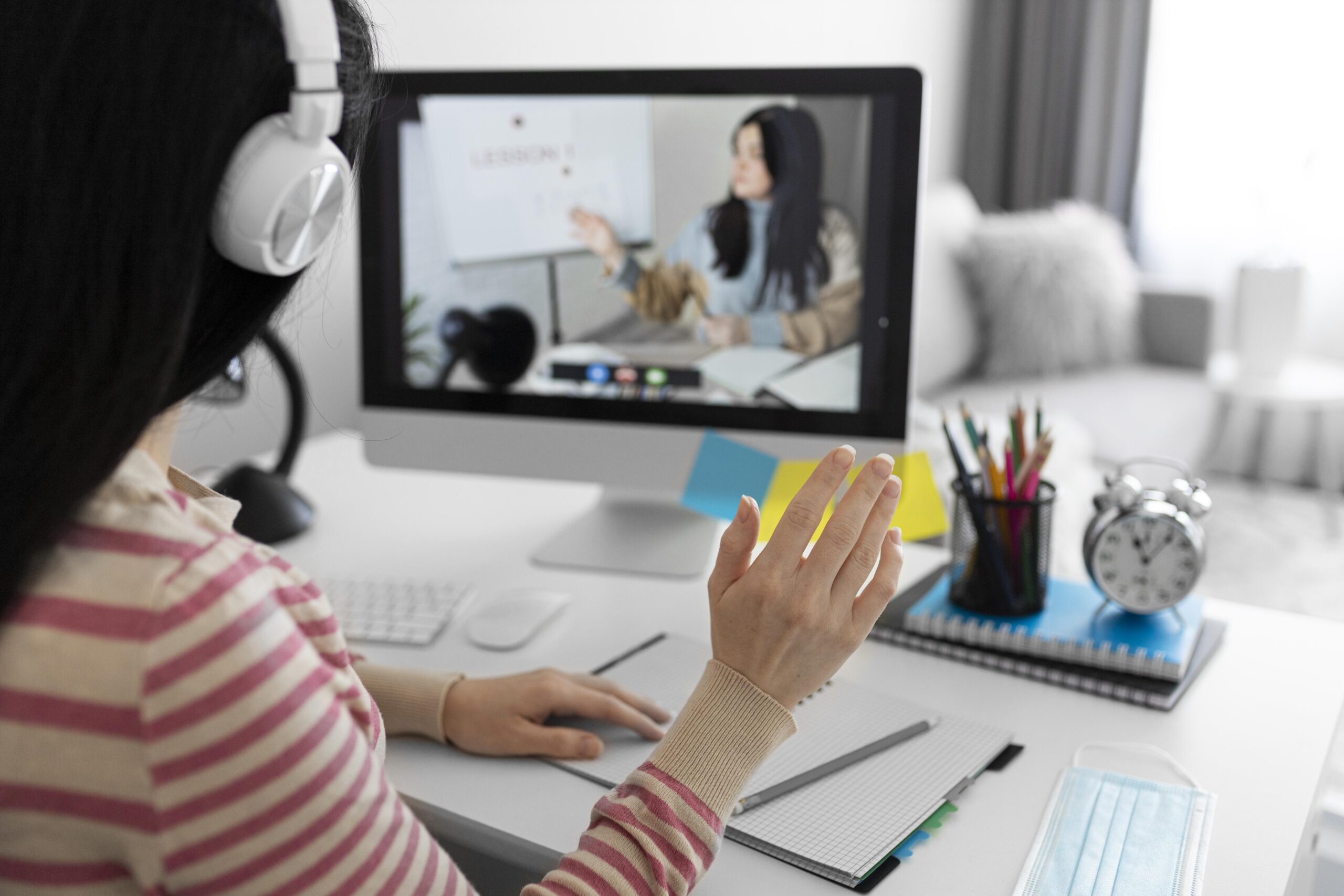 Student attending an online international degree class from home, participating in a live virtual lecture with headphones, computer, and study materials on the desk.