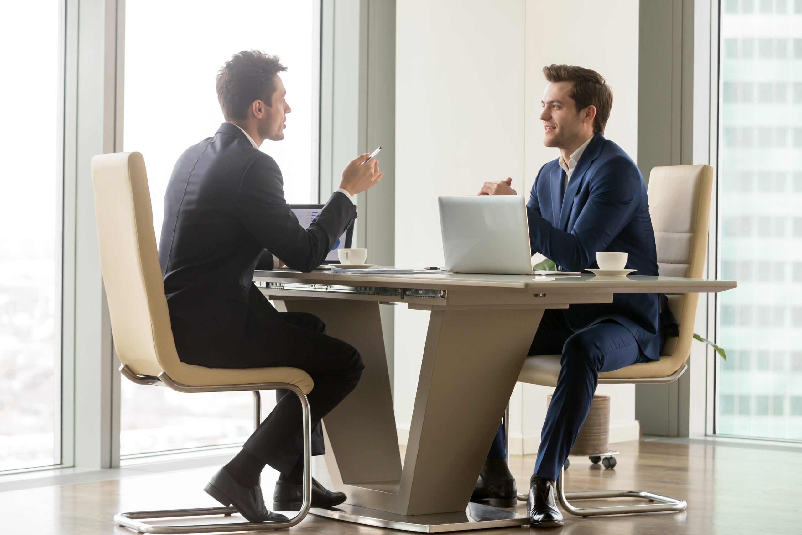 Two business professionals seated across a desk in a modern office, engaged in a collaborative discussion with laptops and coffee cups, representing a professional business meeting or interview.