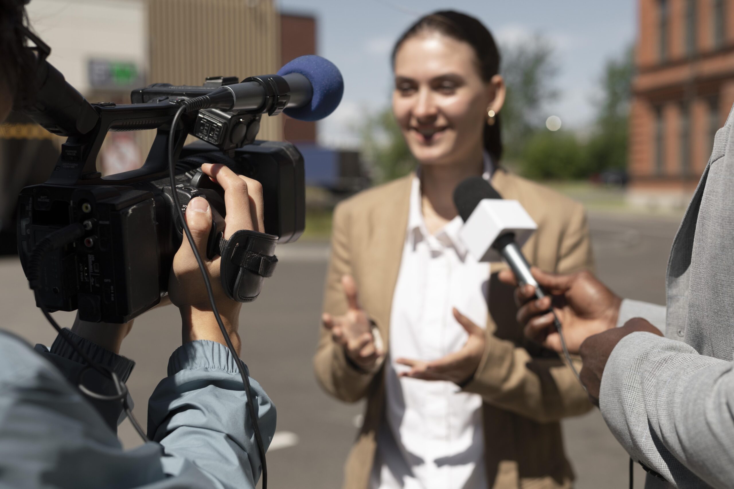 A journalist interviews a woman outdoors while a camera records the conversation and a microphone is held toward her.