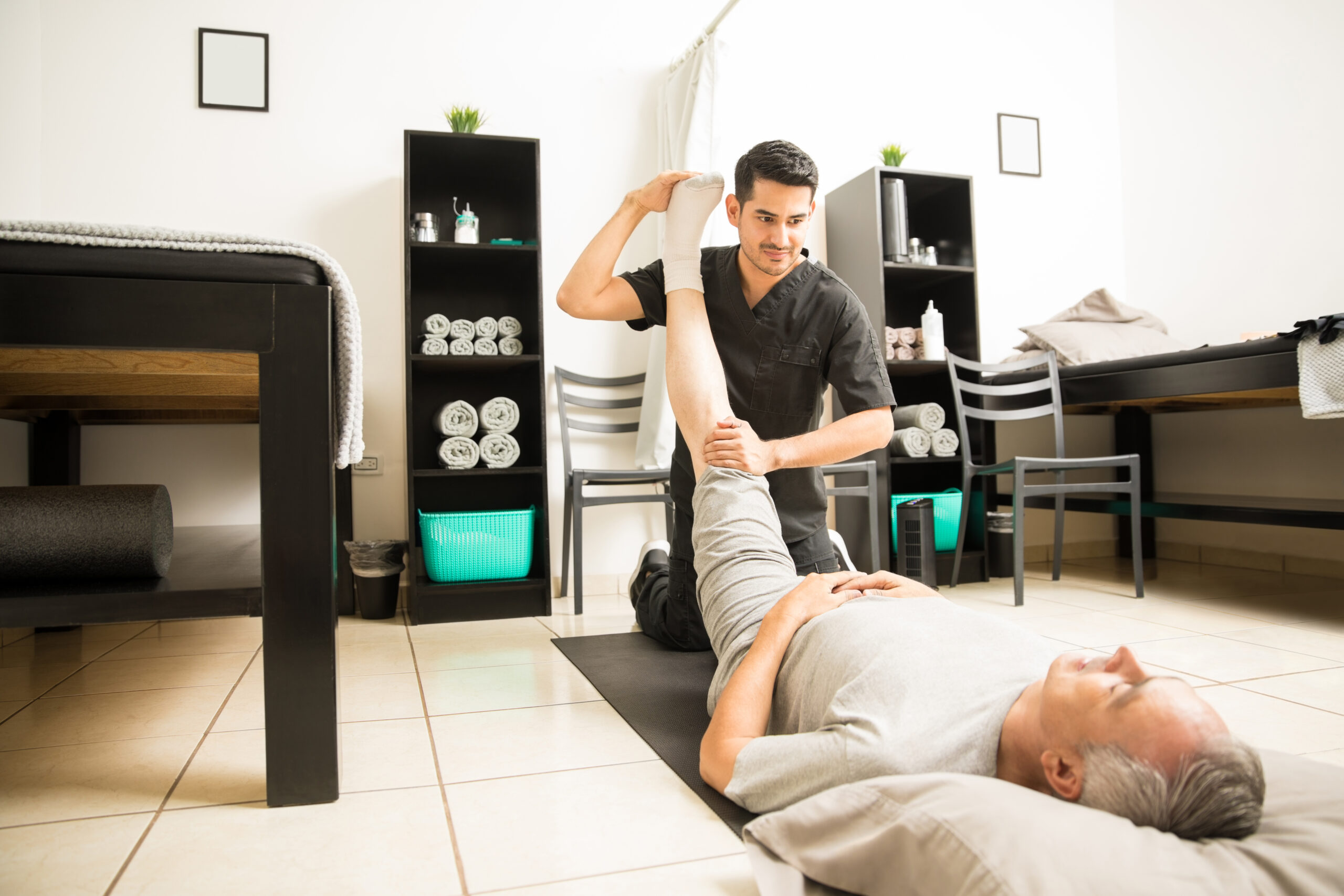 Physiotherapist assisting an older patient with a leg stretching exercise in a bright rehabilitation room.