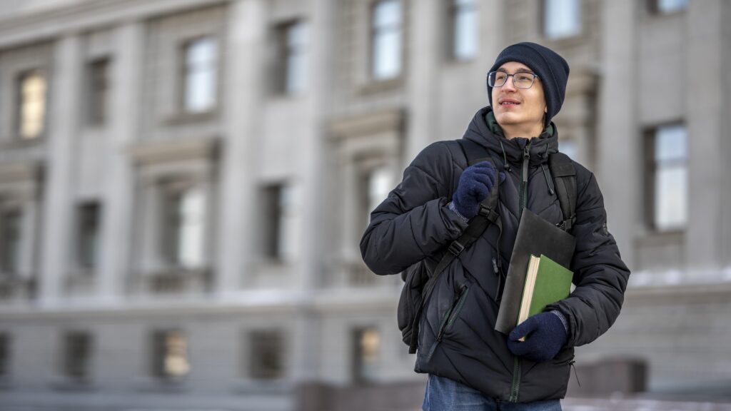 Male student wearing a winter jacket and backpack, carrying books while standing outside a university building.