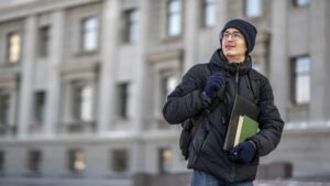 Male student wearing a winter jacket and backpack, carrying books while standing outside a university building.