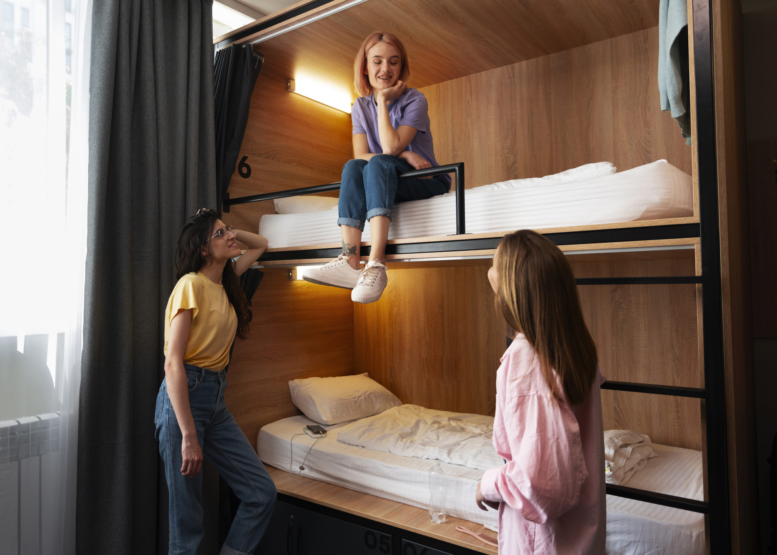 Three young women socializing in a cozy hostel dorm room with bunk beds, smiling and talking together.