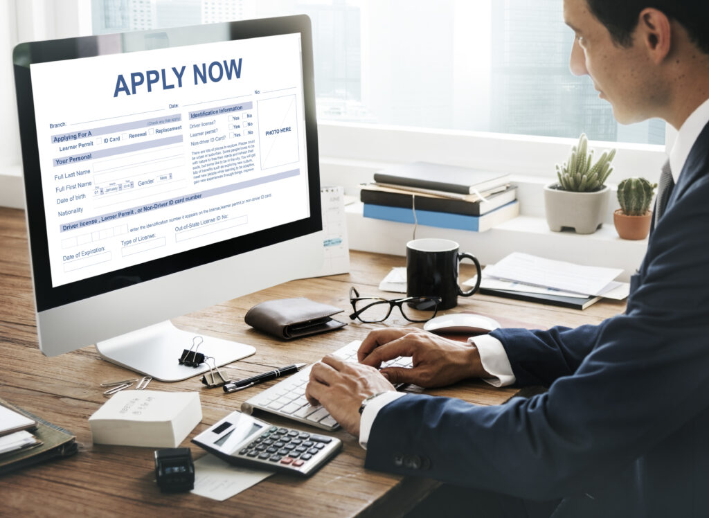 Professional man completing an online application form on a desktop computer at a wooden desk with paperwork, calculator, and personal documents nearby.
