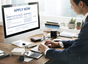 Professional man completing an online application form on a desktop computer at a wooden desk with paperwork, calculator, and personal documents nearby.