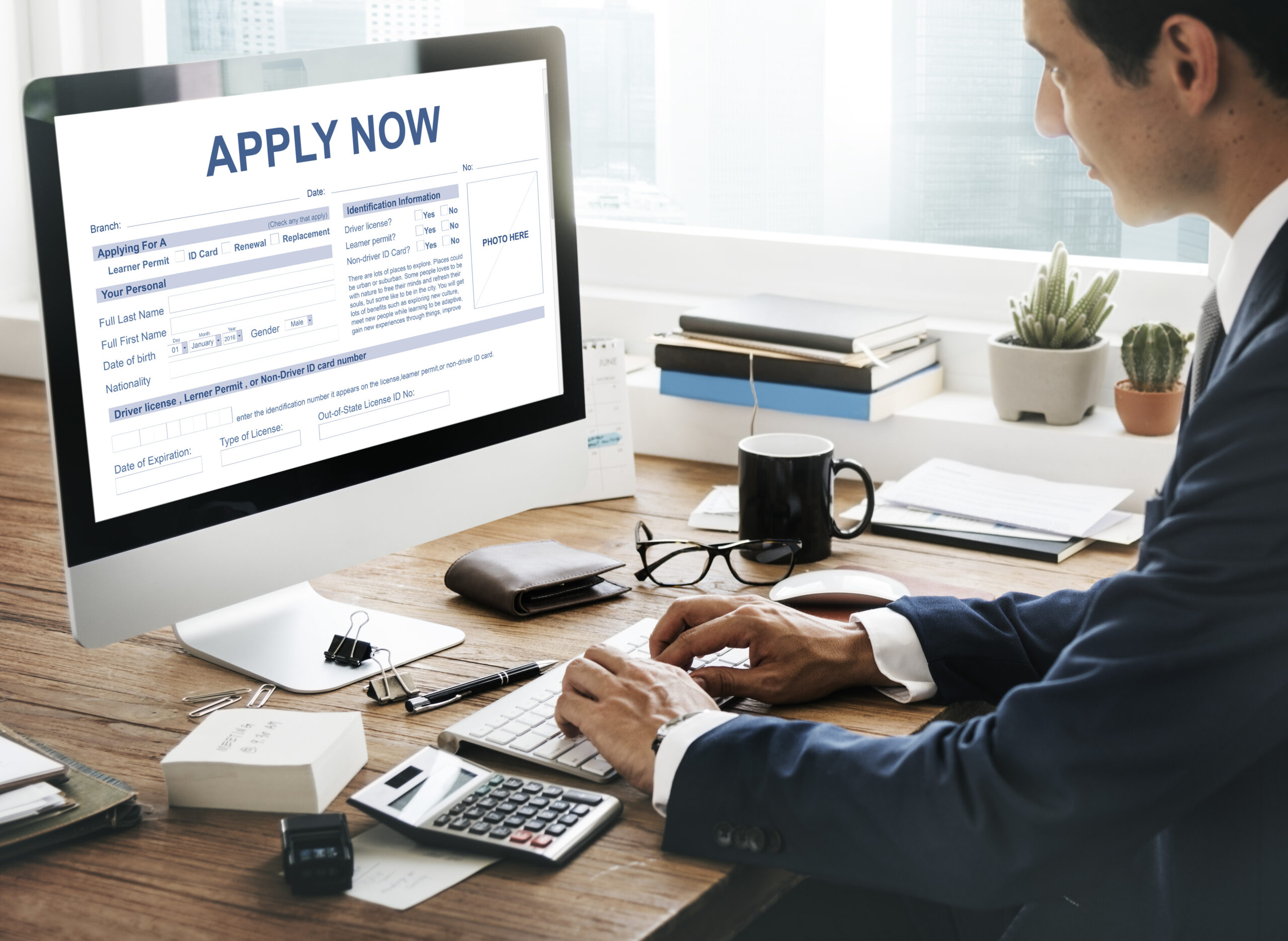 Professional man completing an online application form on a desktop computer at a wooden desk with paperwork, calculator, and personal documents nearby.