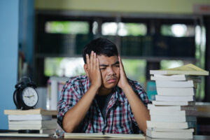 Tired international student studying at a desk surrounded by stacks of books in a library.