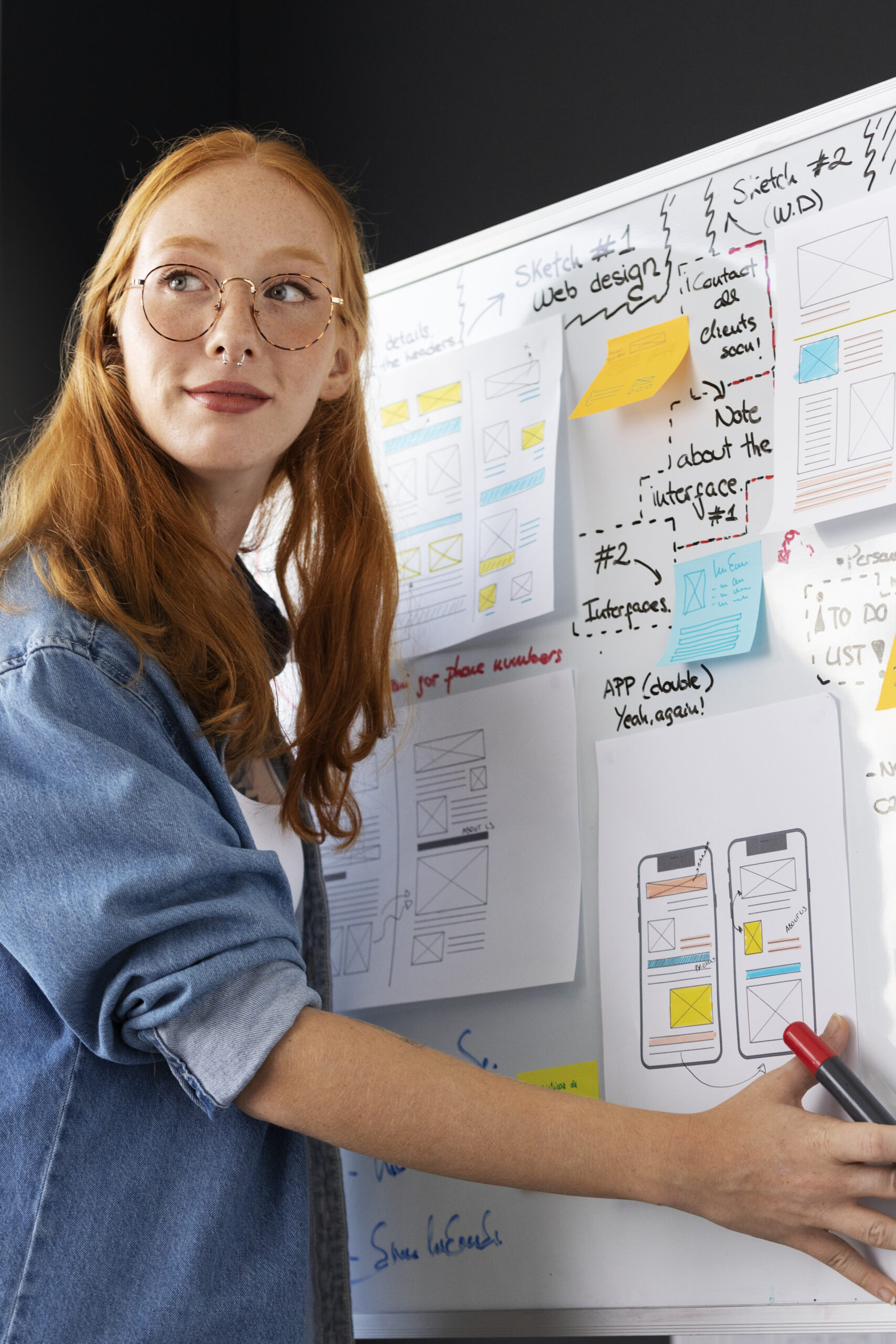 Young web designer presenting website and mobile interface wireframes on a whiteboard in an office.
