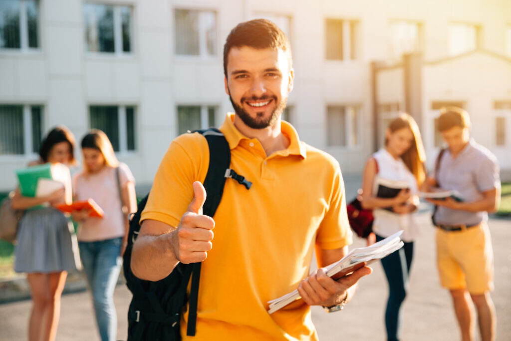Smiling university student with a backpack giving a thumbs-up while holding books on a campus.
